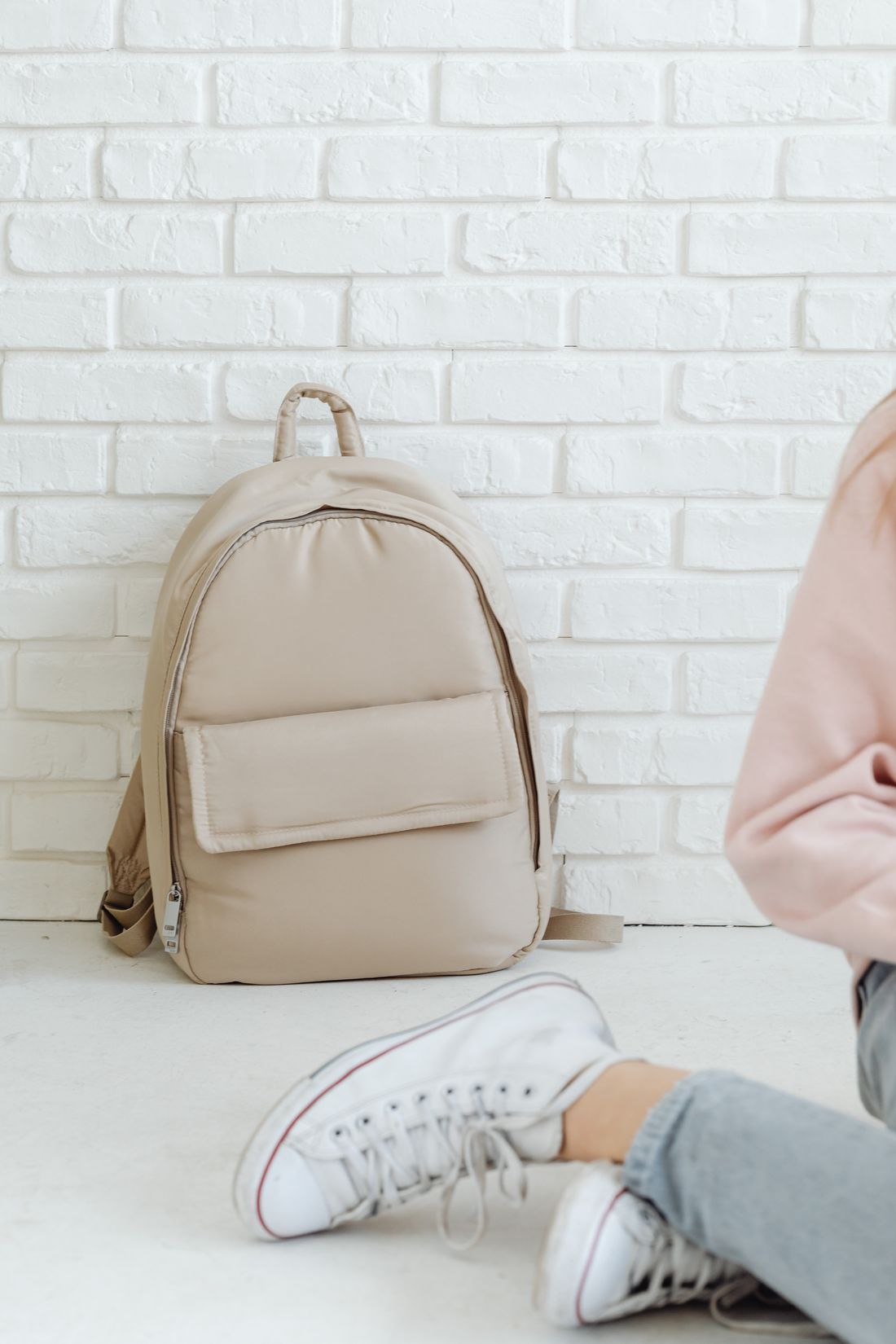 A beige backpack resting against a bright white brick wall.